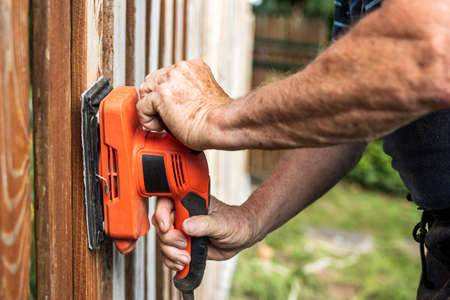Sanding wood. Close-up sander in male hands. Repairing wooden fence at backyardの写真素材