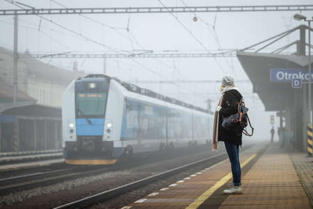 Woman traveler standing on railroad station platform and looking at arriving train from fog. Backpacker travel by trainの写真素材