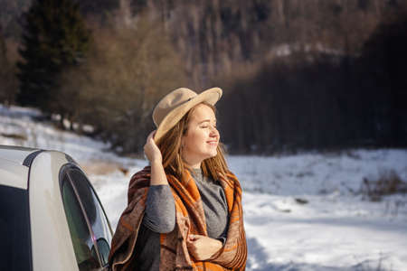 Young woman with hat warming up herself in blanket on sunlight and resting next to car during winter road trip. Enjoyment of sunny day in winter natureの写真素材