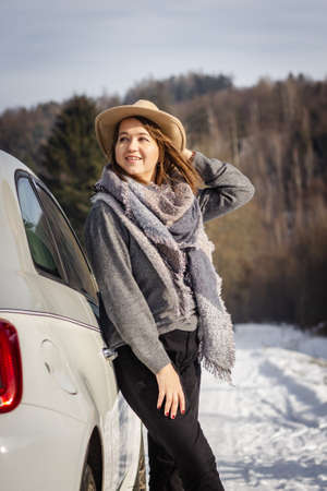 Woman with hat and scarf enjoying sunlight and resting next to car during winter road trip. Young woman is looking to snowy landscapeの写真素材