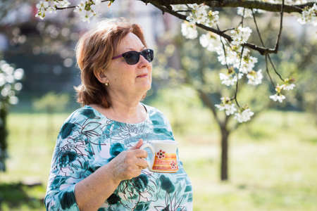 Senior woman drinking coffee in blooming garden. Relaxation outdoors in spring. Female pensioner...
