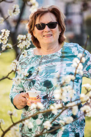 Smiling senior woman drinking coffee in blooming garden. Relaxation and enjoyment outdoors at springtimeの写真素材