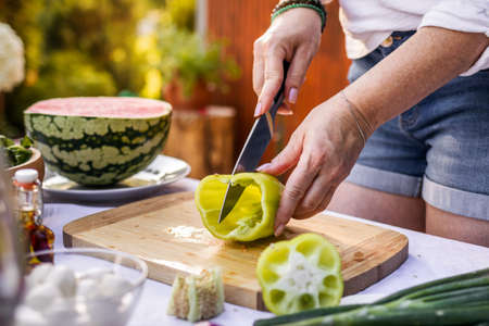 Cutting green bell peppers by kitchen knife. Woman cooking vegetable salad on table outdoors. Preparing healthy foodの写真素材