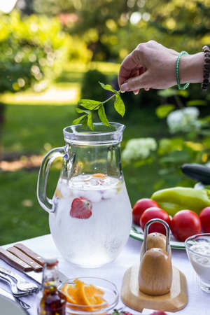 Female hand putting mint herb into cold lemonade in pitcher. Preparing non-alcoholic drink for garden partyの写真素材