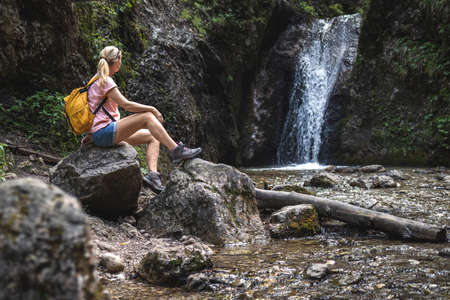 Woman resting during hike in wonderful nature. Hiker sitting on rock near stream with waterfall in forest at natural parkland Mala Fatra, Slovakiaの写真素材
