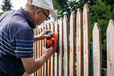 Sanding and preparing wooden fence for painting. Senior man restoring picket fence in gardenの写真素材