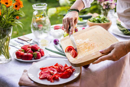 Chopped tomatoes on cutting board. Woman cooking fresh vegetable salad outdoors. Preparing food for garden partyの写真素材