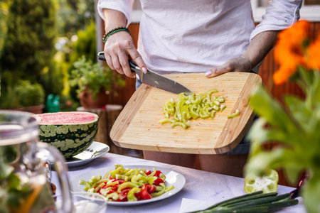 Woman preparing vegetable salad on table. Chopped green bell pepper by kitchen knife on cutting board. Making healthy vegetarian food for summer garden partyの写真素材