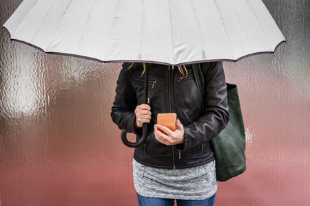 Woman with umbrella holding smart phone and standing in rain in city. Using mobile phoneの写真素材