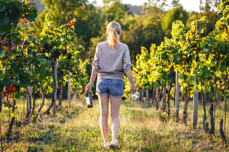 Female vintner holding red wine bottle and drinking glass in vineyard. Woman winetasting outdoorsの写真素材