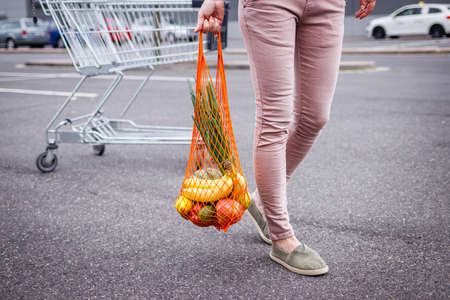 Woman with reusable mesh bag walking from shopping cart in parking lot next to supermarket. Plastic free and zero waste concept. Groceries purchaseの写真素材