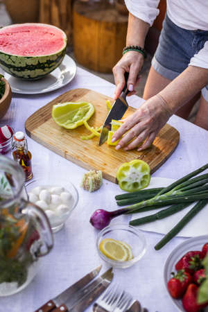 Cutting green bell peppers by kitchen knife. Woman cooking vegetable salad on table outdoors. Preparing healthy foodの写真素材