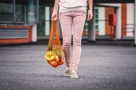 Woman with reusable mesh bag walking at parking lot next to supermarket. Plastic free shopping and...