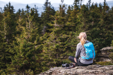 Woman with backpack and camera sitting on rock and enjoying view at forest. Tourist resting and relaxing at mountain peakの写真素材