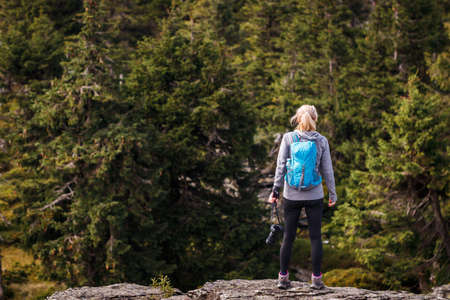 Woman photographer with backpack and camera standing at rocky mountain peak and looking for composition. Hiking and adventure concept. Outdoors active lifestyleの写真素材