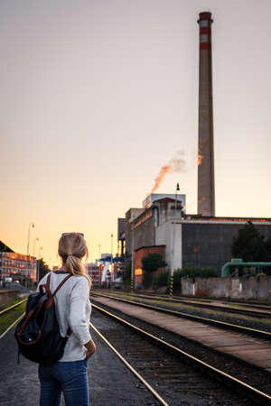Woman waiting for train at railway station during sunset. Industrial area in city. Travel aloneの写真素材