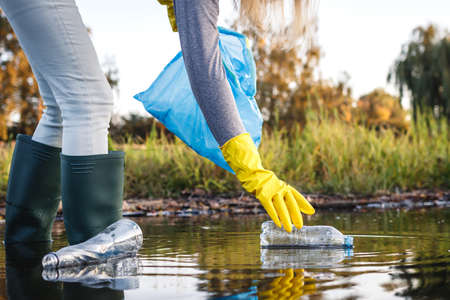 Environmental cleanup. Volunteer picking up plastic bottle from polluted river or lake. Water...