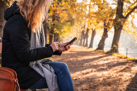 Woman sitting on bench in autumn park and using smart phone. Relaxation and text messaging on mobile phone during city break in fall seasonの写真素材
