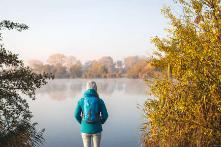 Woman hiking in autumn nature. Female tourist with backpack standing at lake and enjoying view at landscapeの写真素材