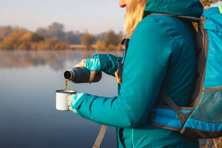 Woman is pouring hot drink from thermos into travel mug. Refreshment during hiking at lake in autumnの写真素材