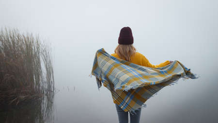 Woman with knit hat is wrapping herself in warm blanket at lake. Female person relaxing in foggy cold autumn morning in nature. Camping outdoorsの写真素材