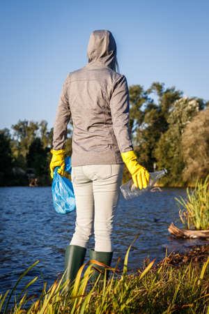 Environmental volunteer with protective glove holding garbage bag and plastic bottle. Female activist standing at lake and cleaning water from plastic wasteの写真素材