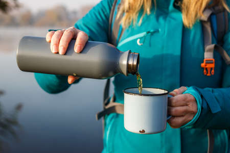 Woman is pouring hot drink from into travel mug. Refreshment during hiking at lake in cold weatherの写真素材