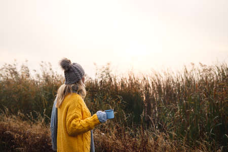Woman relaxing with coffee next to lakeshore at misty cold morning. Camping and hiking outdoors in nature. Blond hair woman wearing knit hat and sweaterの写真素材