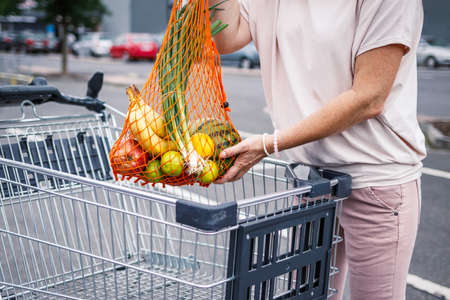 Woman picking up mesh bag with full of fruits from shopping cart at parking lot. Zero waste and plastic free shoppingの写真素材