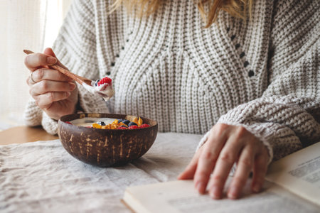 Woman reading book during breakfast at home. Healthy food in coconut bowl. Simple living