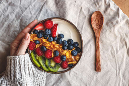 Healthy breakfast in coconut bowl on table with linen tablecloth. Yogurt with corn flakes and blueberry, strawberry, kiwi and raspberry. Womans hand wearing sweater. Vegetarian foodの写真素材