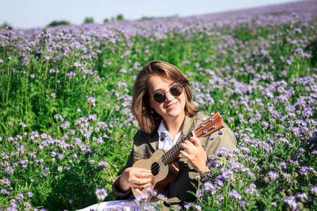 Happy woman playing at ukulele in flowering meadow. Enjoyment of music and spring outdoorsの写真素材