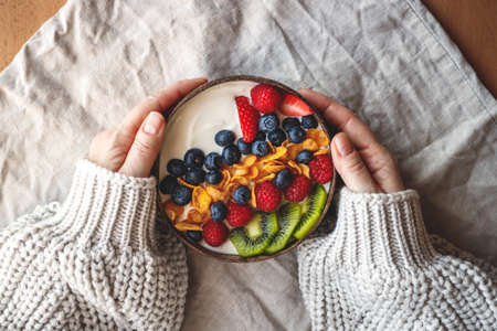 Healthy breakfast in coconut bowl on table with linen tablecloth. Yogurt with corn flakes and blueberry, strawberry, kiwi and raspberry. Womans hand wearing sweater. Vegetarian foodの写真素材