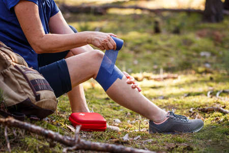Injured hiker putting elastic bandage to her knee. Injury during hiking. First aid kit. Knee joint pain and tendon problems during adventure in natureの写真素材