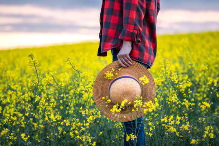 Farmer standing in blooming oilseed field. Woman holding straw hat in rapeseed plants. Farming and agriculture conceptの写真素材