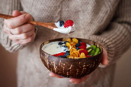 Woman eating healthy breakfast from coconut bowl. Yogurt with corn flakes and berry fruit. Eco friendly and zero waste tablewareの写真素材