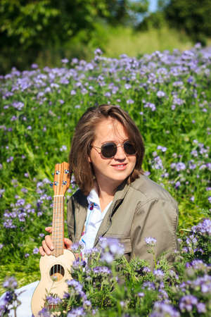 Candid portrait of happy young smiling woman with ukulele enjoying sunlight in flowering meadow at springtimeの写真素材