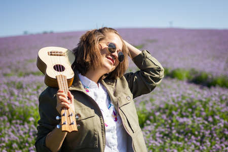 Portrait of happy young smiling woman with ukulele enjoying sunlight in flowering meadow at springの写真素材