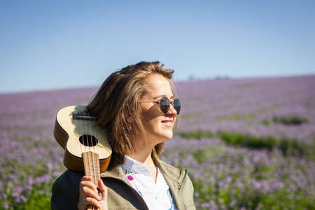 Candid portrait of happy young smiling woman with ukulele enjoying sunlight in flowering meadow at springの写真素材