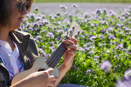Happy woman playing at ukulele in flowering meadow. Enjoyment of music and spring outdoorsの写真素材
