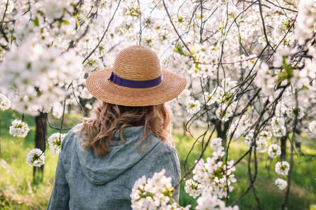 Woman wearing linen shirt and straw hat in blooming orchard. Enjoyment of springtime outdoors. Boho fashion styleの写真素材