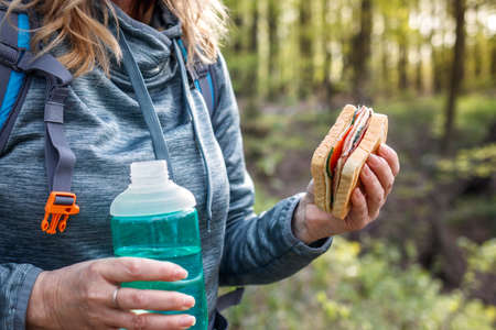 Woman tourist eating sandwich and drinking water outdoors. Refreshment during hiking in forestの写真素材