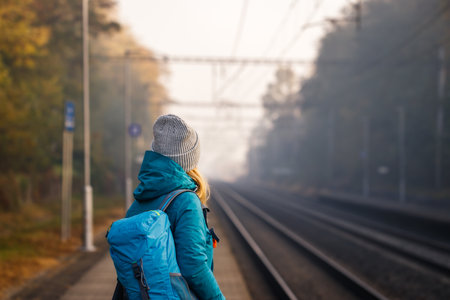 Female tourist with backpack waiting for train on empty railroad station alone. Woman travel at fall...