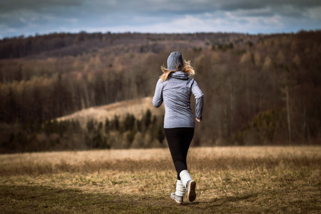 Woman running in autumn nature. Fitness activity outdoors. Female athlete wearing knit hat, hoodie, leggings and leg warmers during sports trainingの写真素材