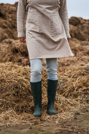 Woman farmer with apron and rubber boots standing next to organic manure pile. Autumn agricultural activityの写真素材