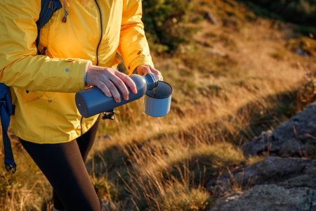 Hiker is pouring hot drink from thermos into travel mug in mountains. Refreshment during trekking in nature. Woman wearing sports clothing and using hiking equipmentの写真素材