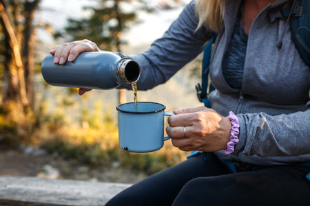 Woman pouring tea drink into travel mug. Refreshment during hiking in the forestの写真素材