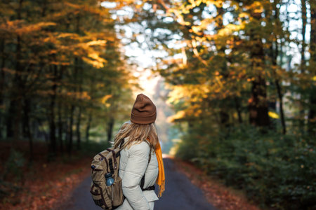 Woman with backpack, jacket, scarf and knit hat hiking on road in autumn forest. Fall season adventure in natureの写真素材