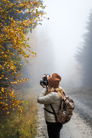 Woman with camera taking picture of autumn leaf. Tourist hiking in misty forest. Landscape photographer with backpack enjoying nature in fall seasonの写真素材