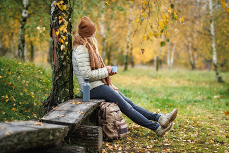 Relaxation during autumn hiking. Woman drinks hot coffee and resting on bench in forest with birch treesの写真素材
